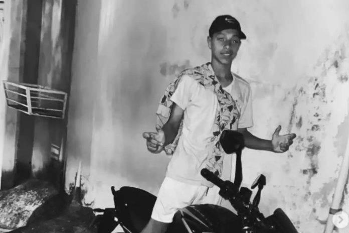 expresso-439a75bd2a02f219a5319485 Fotografia em preto e branco de um adolescente posando em frente a uma parede, com uma motocicleta visível parcialmente. Imagem vinculada a redes sociais, usada para identificação do jovem falecido e tratada como foto de arquivo.