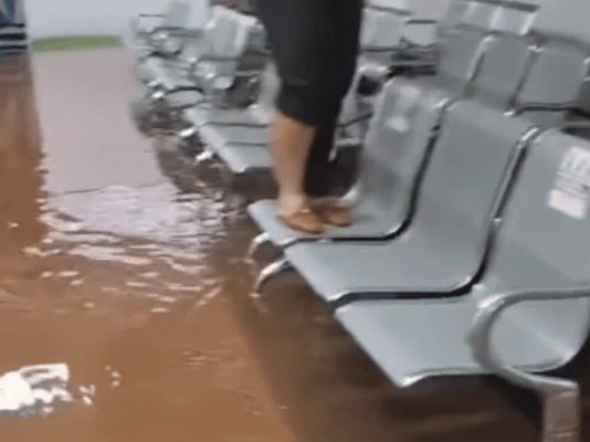Chuva Intensa Causa Inundações e Danos em Paulicéia, SP Área interna alagada na unidade de saúde em Paulicéia durante forte tempestade, com água cobrindo o chão e pessoa em cima das cadeiras para evitar contato com a água.