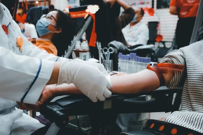Pessoa doando sangue em ambiente coletivo, mostrando preparação para coleta com profissional de saúde usando luvas.