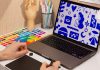 Carapicuíba lança oficina gratuita de design gráfico com inscrições online Mãos utilizando mesa digitalizadora gráfica, computador com ilustrações e paleta de cores, em ambiente de trabalho de design gráfico.