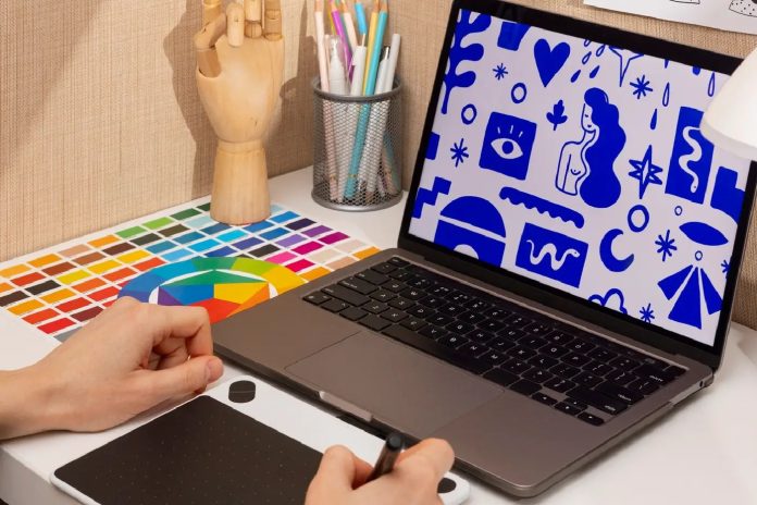 Mãos utilizando mesa digitalizadora gráfica, computador com ilustrações e paleta de cores, em ambiente de trabalho de design gráfico.