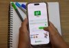 Famílias de Osasco Ganham Aplicativo para Acompanhar Vida Escolar dos Alunos Pessoa segurando um celular com a tela de login do aplicativo Portal do Aluno da Prefeitura de Osasco, sobre uma mesa com caderno e canetas.
