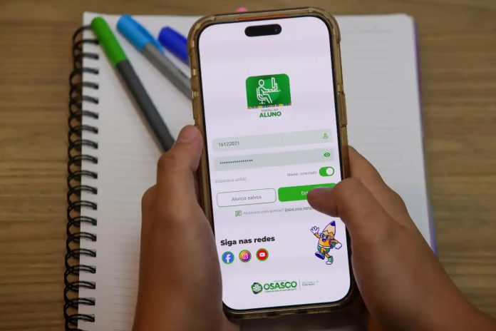 Pessoa segurando um celular com a tela de login do aplicativo Portal do Aluno da Prefeitura de Osasco, sobre uma mesa com caderno e canetas.